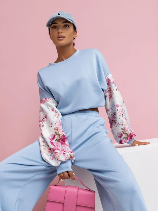 Zdjęcie przedstawia komplet damski dresowy w kolorze niebieskim. Komplet składa się z bluzy i spodni. Bluza bez kaptura wkłada przez głowę z bufiastymi rękawami z efektownym kwiatowym wzorem. Spodnie z długimi i szerokimi nogawkami.